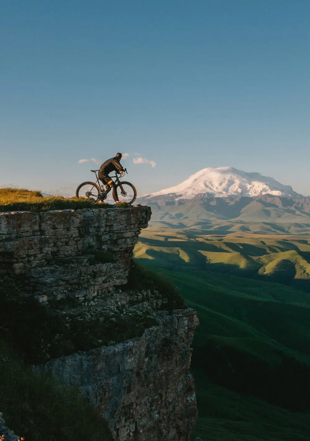 rider stopped with a bike on a grassy cliff edge overlooking green valleys and a snowy peak, image for funny biking puns