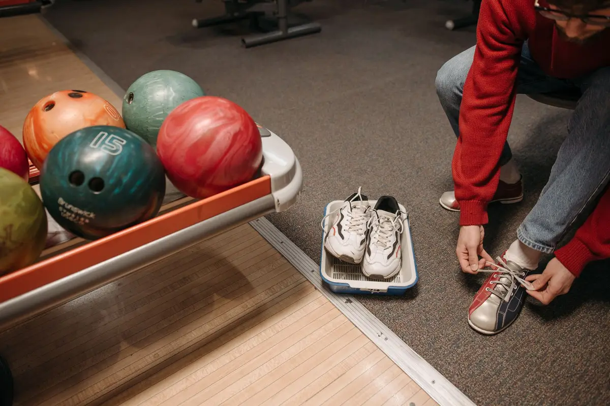 man tying rental bowling shoes beside colorful bowling balls, bowling jokes one liners