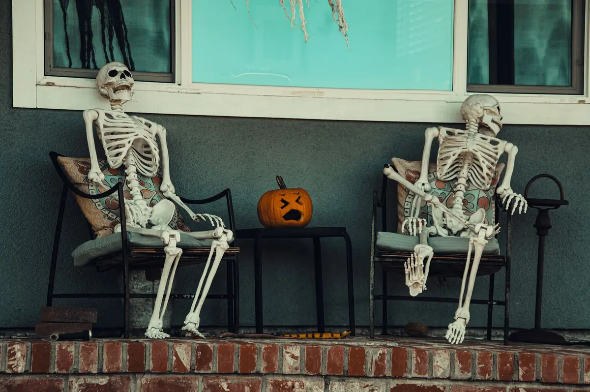 two skeletons sitting on porch chairs beside a carved pumpkin, skeleton jokes