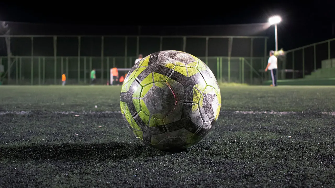 a football resting on an artificial pitch under floodlights at night, image for everton matchday captions