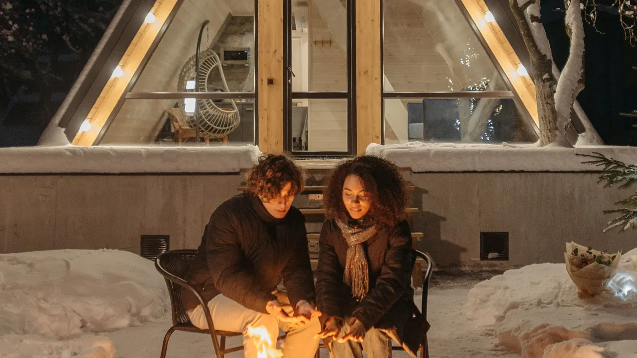 two people sitting by a campfire in front of a lit a-frame cabin in the snow, image for deer camp jokes