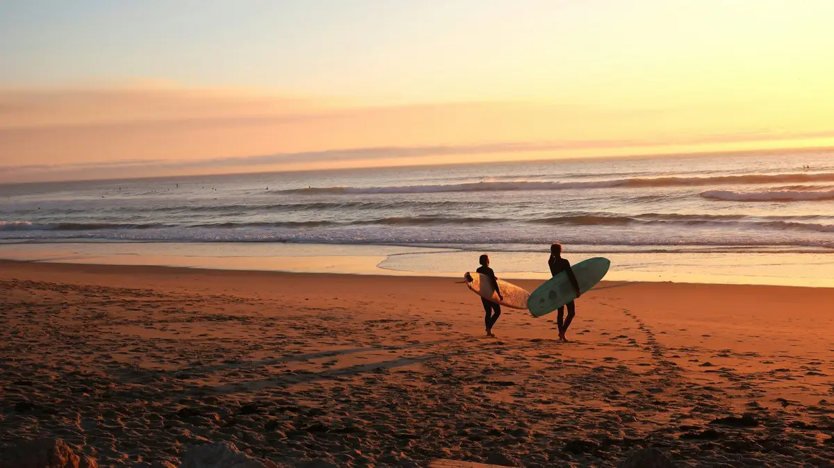 two surfers carrying boards toward the water on a beach at sunset, image for surfing pick up lines