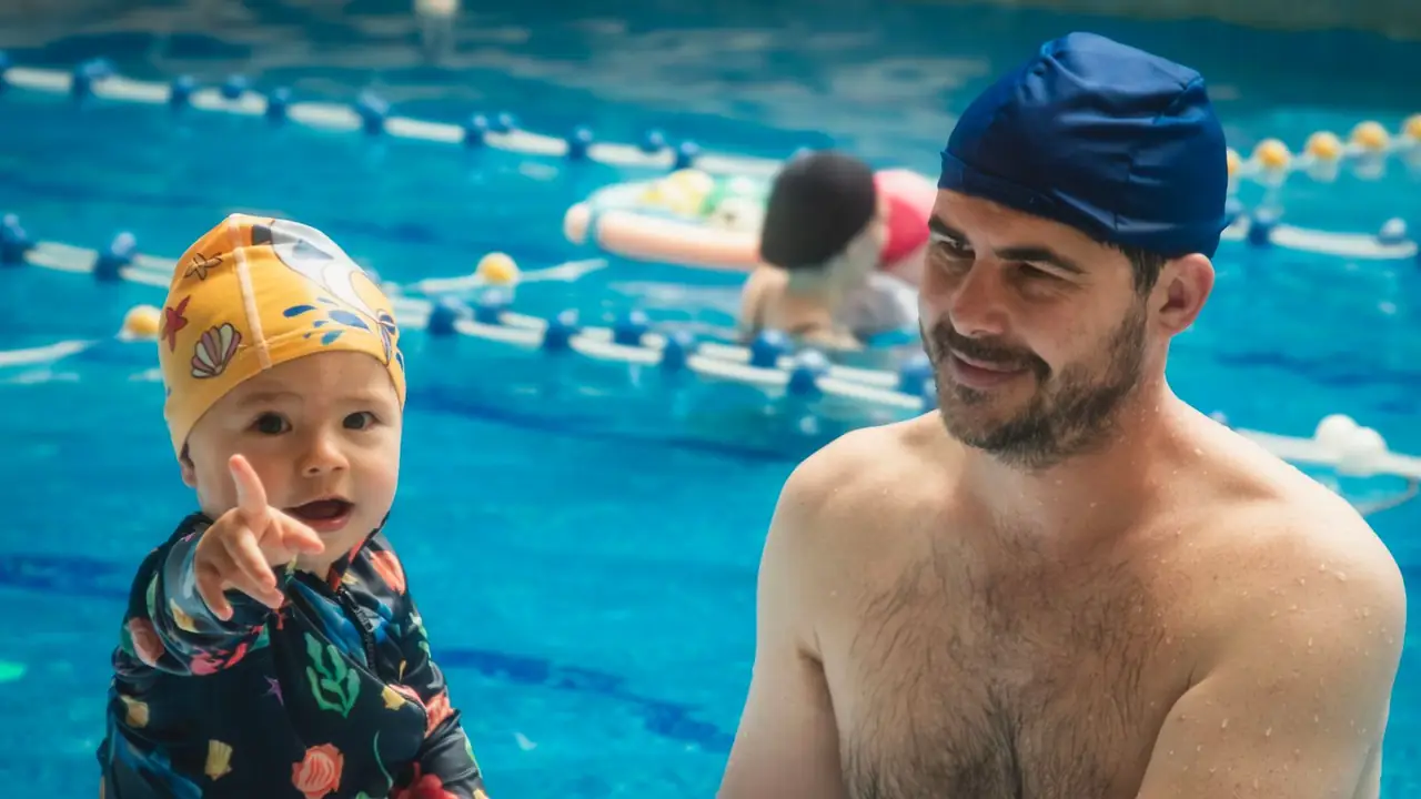 a dad in a swim cap smiling with a baby in the pool, image for swimming dad jokes at the shallow end