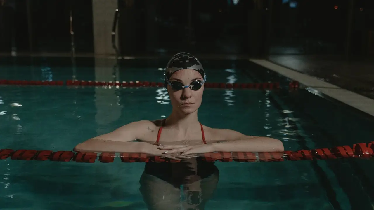 a swimmer in a cap and goggles resting on a lane rope in a dim indoor pool, image for swim meet dad jokes